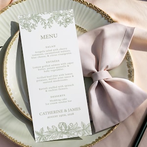 May include: A wedding menu card with a floral design in green ink, featuring a list of dishes. The card is placed on a decorative plate with a gold rim, next to a folded pink napkin. The menu includes salad, entrees, and desserts.