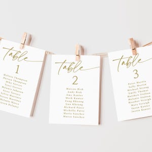 Pu&ograve; includere: Tre segnaposto bianchi con calligrafia dorata, appesi a una corda con mollette di legno. Le carte mostrano la parola "Table" e un numero, pi&ugrave; i nomi degli ospiti. Ideali per matrimoni o eventi.