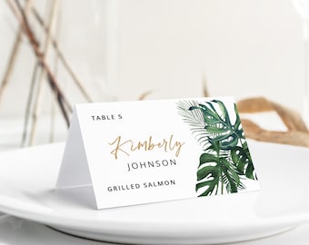 May include: A white folded place card featuring the text TABLE 5, Kimberly, JOHNSON, and GRILLED SALMON. The card is decorated with a watercolor illustration of green monstera and palm leaves on the right side, with gold script lettering.