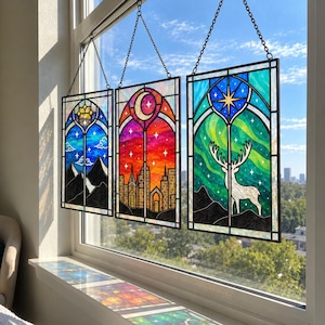 Puede incluir: Tres paneles rectangulares de vidrieras colgados en una ventana. El panel izquierdo muestra montañas azules y un sol. El panel central representa el horizonte de una ciudad bajo una luna creciente. El panel derecho muestra un ciervo blanco y auroras.