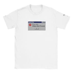 Può includere: Una t-shirt bianca a maniche corte con una stampa grafica che raffigura una finestra grigia di errore di sistema del computer. La finestra mostra il testo 'System Error' in alto e 'Error 404: Vibes Not Found' con un'icona 'X' rossa.