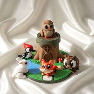Puede incluir: Un expositor circular con un tronco de árbol marrón central con puerta y un búho posado encima. Alrededor de la base hay un conejo blanco, un pájaro carpintero blanco y negro, un zorro naranja y un oso marrón sobre una base verde.