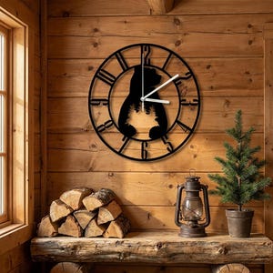 Puede incluir: Un reloj de pared circular de metal negro con una silueta recortada de un oso que contiene una escena de bosque en su interior. El reloj presenta números negros grandes y llamativos en el borde y dos manecillas blancas que marcan la hora.