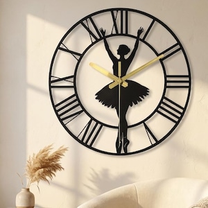 May include: A circular wall clock featuring a black metal frame with Roman numerals and a silhouette of a ballerina in the centre. The clock has two gold-coloured hands positioned against the open-work design of the dancer in a tutu.