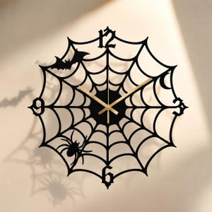 May include: A black wall clock featuring a spiderweb design with a silhouette of a bat and a spider integrated into the frame. The clock has gold-colored hands and black numerals at the 3, 6, 9, and 12 positions, creating a circular shape.