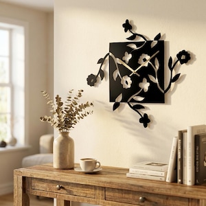 May include: A square black wall clock featuring a cutout floral and vine design that extends beyond the edges of the square frame. The clock has two thin gold-colored hands centered on the black surface, displaying a botanical motif.