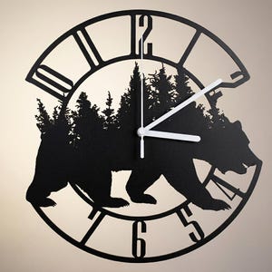 May include: A circular black wall clock featuring a cutout silhouette of a bear walking through a forest of pine trees. The clock face has large, bold, open-style numerals around the perimeter and thin white hour, minute, and second hands.