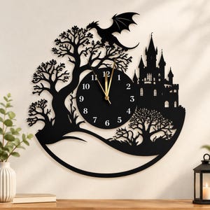 May include: A black wall clock featuring a circular face with white numbers and gold hands. The clock is surrounded by a laser-cut silhouette design depicting a large castle, a flying dragon, and two bare trees, all integrated into a circular frame.