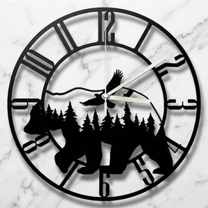 May include: A circular black wall clock featuring a cutout silhouette of a bear walking through a forest with a flying eagle above. The clock face displays large Roman numerals around the perimeter and includes thin silver-colored clock hands.