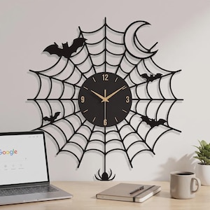 May include: A black wall clock featuring a spiderweb design with a crescent moon and four bat silhouettes. The circular clock face has gold-colored hands and numerals at the 3, 6, 9, and 12 positions. A small spider hangs from the bottom of the web.