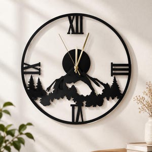 May include: A circular black wall clock featuring a mountain and forest silhouette design. The clock face displays Roman numerals at the twelve, three, six, and nine o'clock positions, with thin gold-colored hour and minute hands at the center.