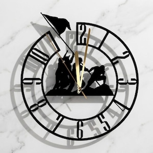 May include: A circular wall clock featuring a black silhouette of soldiers raising a flag. The clock face has large, cut-out black numerals from one to twelve. Two thin, gold-coloured metal hands indicate the time, centred over the central scene.