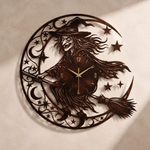 May include: A circular wall clock featuring a dark brown, laser-cut metal design of a witch flying on a broomstick in front of a crescent moon. The clock face is centered on the witch's body, displaying gold-colored hands and numbers 3, 9, and 12.