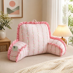 May include: A pink and white striped reading pillow featuring a floral pattern, ruffled edges, and a small side pocket. The backrest cushion has a handle at the top and is designed with a structured shape to provide support while sitting upright.