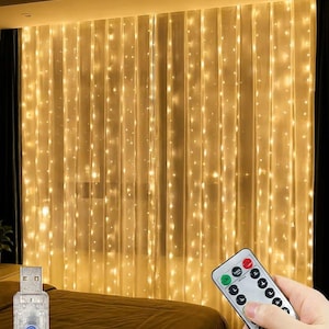 May include: A curtain of warm white LED string lights hanging vertically, featuring a USB connector with a blue button and a white remote control with multiple buttons for adjusting light modes and brightness settings for the illuminated display.