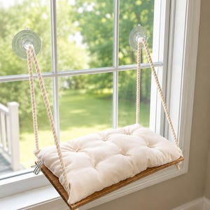 May include: A rectangular wooden platform suspended by two thick twisted ropes attached to the window glass with clear suction cups. A tufted, off-white rectangular cushion rests on the wooden base, creating a hanging shelf structure.