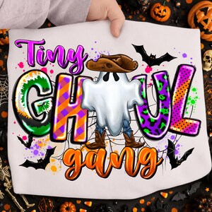 Può includere: Una t-shirt bianca con una grafica colorata e la scritta Tiny Ghoul Gang. Il design include un fantasma bianco con cappello e stivali da cowboy marroni, circondato da pipistrelli neri, ragnatele e motivi vivaci viola, arancioni e verdi.