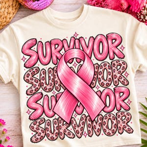 Op de afbeelding: Een crèmekleurig t-shirt met een grote roze bewustwordingslint gecentreerd over het woord SURVIVOR dat meerdere keren in een gestapeld ontwerp staat. De tekst wisselt af tussen een effen roze penseelstijl en een roze luipaardprint.