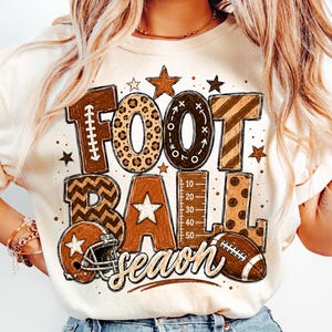 May include: A cream-coloured t-shirt featuring a large graphic with the text FOOTBALL SEASON. The letters are decorated with brown patterns including leopard print, chevron, and stripes. A football helmet and a football are also illustrated.