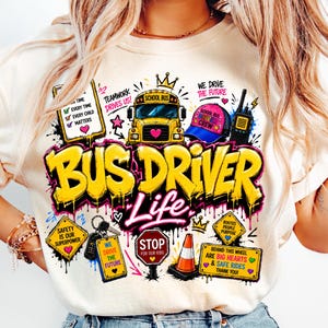 Può includere: Una t-shirt color crema con una stampa grafica colorata raffigurante uno scuolabus giallo, un berretto rosa e blu, un walkie-talkie, un segnale di stop e coni stradali. Il testo recita Bus Driver Life, Teamwork Drives Us, We Drive The Future, Safety Is Our Superpower e altre frasi.