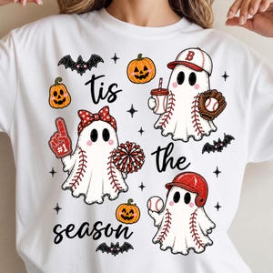 Könnte beinhalten: Ein weißes T-Shirt mit einem Grafikdruck und dem Text 'tis the season'. Das Design zeigt drei Geister mit Baseball-Nähten, einen Cheerleader-Geist mit roter Schleife und Pompons, Baseball-Ausrüstung, Fledermäuse und Kürbisse.