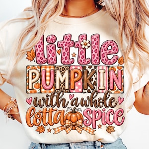 May include: A cream-colored t-shirt featuring a graphic design with the text "little PUMPKIN with a whole lotta spice". The letters are filled with pink leopard print, orange plaid, and brown patterns, accented by small pumpkins, leaves, and stars.