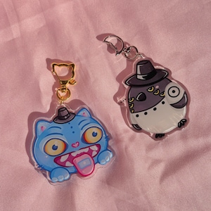 May include: Two acrylic keychains featuring cartoon characters. One is a blue cat-like creature with a top hat, wide eyes, and a pink tongue sticking out, attached to a gold cat-shaped clasp. The other is a grey and white round creature with a top hat.