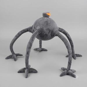Peut inclure: Une peluche en forme de créature avec un corps rond gris foncé et quatre longues pattes segmentées se terminant par des pieds à trois doigts. La créature présente une petite protubérance sur le dessus avec un accent orange vif.