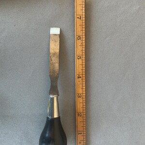 May include: A metal chisel with a black, pear-shaped wooden handle and a silver-colored metal ferrule. The blade is stamped with the word SARGENT. A wooden ruler is placed alongside the tool, showing a total length of approximately 8.5 inches.