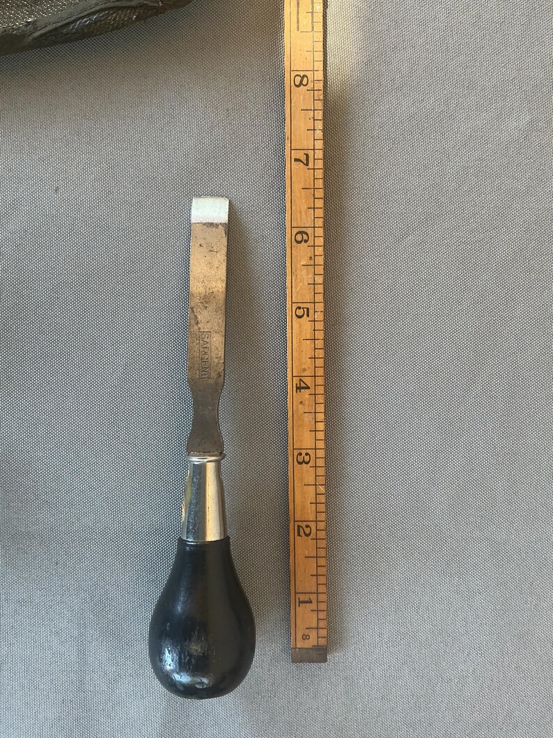 May include: A metal chisel with a black, pear-shaped wooden handle and a silver-colored metal ferrule. The blade is stamped with the word SARGENT. A wooden ruler is placed alongside the tool, showing a total length of approximately 8.5 inches.