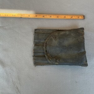 May include: A dark gray, rectangular, textured pouch with a rounded flap and three visible metal rivets. A yellow wooden ruler is positioned above the pouch, showing a width of approximately 5 inches for the item.