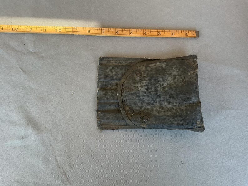 May include: A dark gray, rectangular, textured pouch with a rounded flap and three visible metal rivets. A yellow wooden ruler is positioned above the pouch, showing a width of approximately 5 inches for the item.