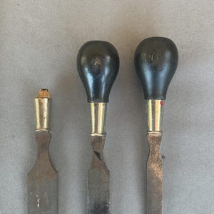 May include: Three metal chisels with flat blades and rounded black wooden handles. The two larger tools feature bulbous black handles with silver-toned metal ferrules, while the smallest tool has a broken wooden handle and a silver-toned ferrule.