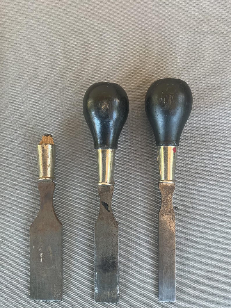 May include: Three metal chisels with flat blades and rounded black wooden handles. The two larger tools feature bulbous black handles with silver-toned metal ferrules, while the smallest tool has a broken wooden handle and a silver-toned ferrule.