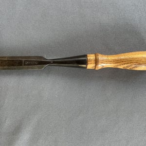May include: A steel wood chisel with a light brown wooden handle and a dark metal ferrule, positioned horizontally next to a yellow wooden ruler that shows the tool is approximately 6 inches long from the tip to the end of the handle.