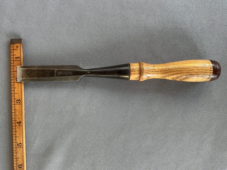 May include: A steel wood chisel with a light brown wooden handle and a dark metal ferrule, positioned horizontally next to a yellow wooden ruler that shows the tool is approximately 6 inches long from the tip to the end of the handle.