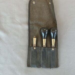 May include: A dark brown canvas tool roll containing three handheld tools. Two tools feature rounded black handles with brass-colored ferrules, while the third tool is a slender metallic cylinder. The roll has a flap secured with metal snaps.