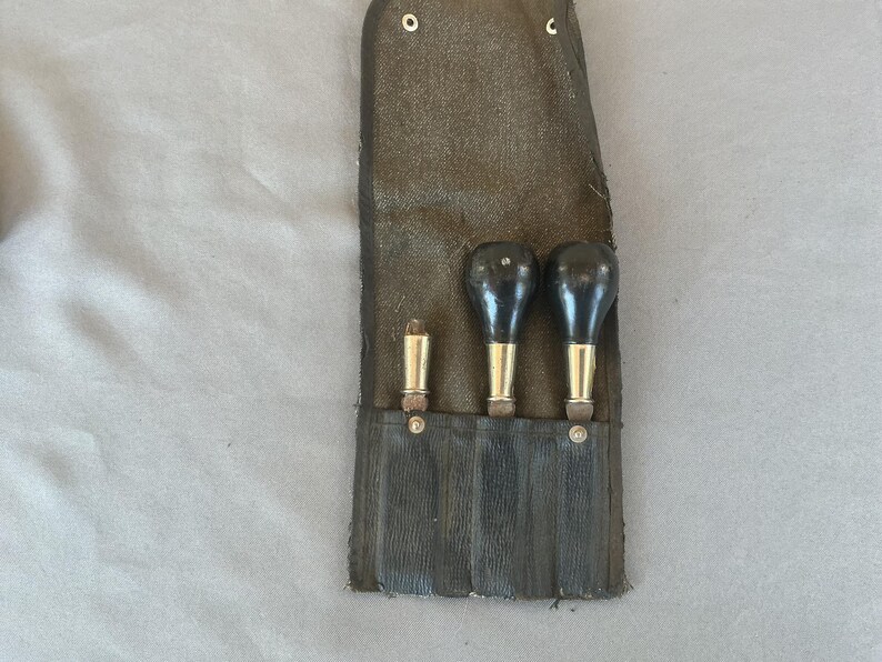 May include: A dark brown canvas tool roll containing three handheld tools. Two tools feature rounded black handles with brass-colored ferrules, while the third tool is a slender metallic cylinder. The roll has a flap secured with metal snaps.