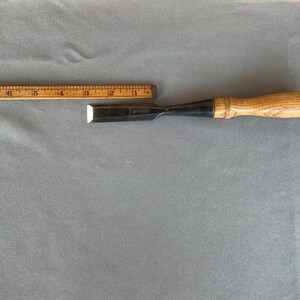 May include: A wood chisel with a metallic blade and a light brown wooden handle, positioned alongside a wooden ruler that shows the tool measures approximately 8 inches in total length from the tip of the blade to the end of the handle.