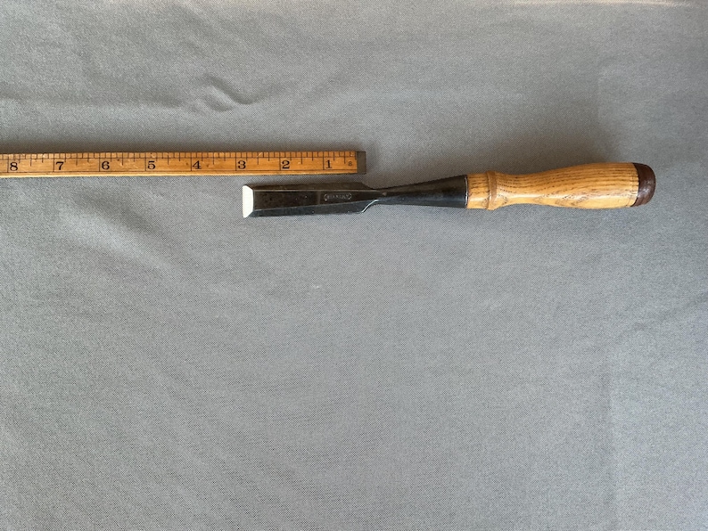 May include: A wood chisel with a metallic blade and a light brown wooden handle, positioned alongside a wooden ruler that shows the tool measures approximately 8 inches in total length from the tip of the blade to the end of the handle.