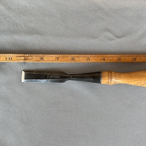 May include: A steel wood chisel with a light-colored wooden handle positioned next to a yellow wooden ruler. The ruler shows the chisel blade is approximately 4 inches long, with the total tool length measuring about 9.5 inches in total.