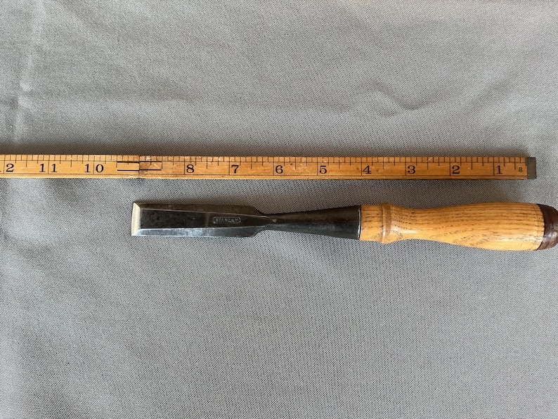 May include: A steel wood chisel with a light-colored wooden handle positioned next to a yellow wooden ruler. The ruler shows the chisel blade is approximately 4 inches long, with the total tool length measuring about 9.5 inches in total.