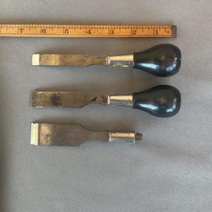 May include: Three metal chisels with silver-toned ferrules and black handles, arranged horizontally below a wooden ruler that shows measurements in inches. The top two chisels have rounded handles, while the bottom chisel lacks a handle.