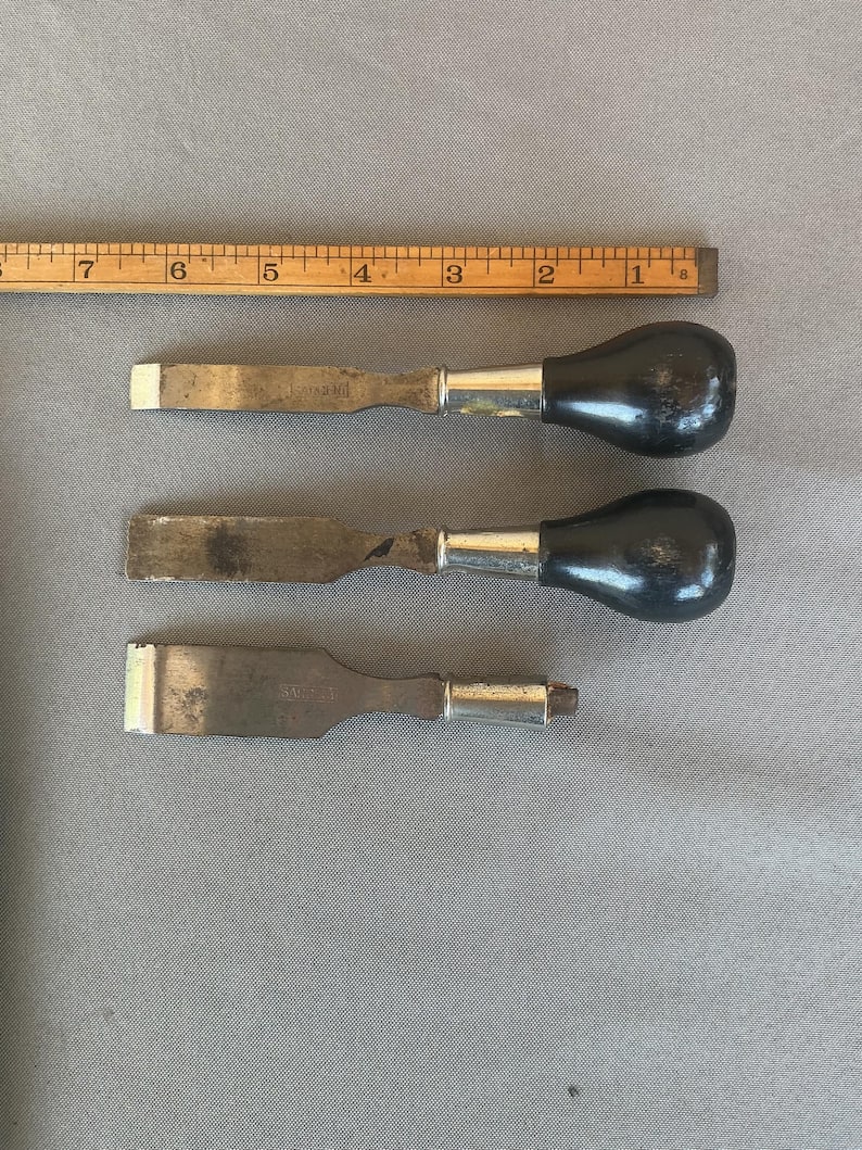 May include: Three metal chisels with silver-toned ferrules and black handles, arranged horizontally below a wooden ruler that shows measurements in inches. The top two chisels have rounded handles, while the bottom chisel lacks a handle.