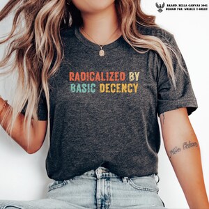 May include: A dark heather grey short-sleeve t-shirt featuring the text "RADICALIZED BY BASIC DECENCY" in a distressed, multi-coloured retro font. The shirt has a crew neckline and is paired with light blue denim jeans.