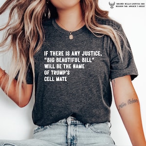 May include: A dark heather gray short-sleeve t-shirt featuring white text that reads: IF THERE IS ANY JUSTICE, “BIG BEAUTIFUL BILL” WILL BE THE NAME OF TRUMP’S CELL MATE. The shirt has a crew neckline and is worn with light blue denim jeans.