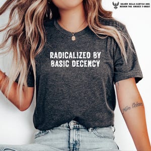 Op de afbeelding: Een donkergrijs gemêleerd T-shirt met korte mouwen en de witte tekst "RADICALIZED BY BASIC DECENCY" in een verweerd lettertype op de borst. Het shirt heeft een ronde hals en is gemaakt van een zachte, gemêleerde stof.