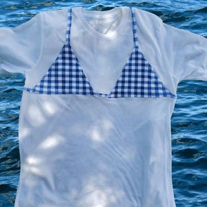 May include: A white short-sleeved t-shirt featuring a blue and white gingham patterned bikini top overlay. The garment includes thin straps and triangular cups stitched onto the front. The text @mendedtide is visible in the top right corner.