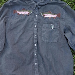 May include: A charcoal gray long-sleeve button-down shirt featuring two embroidered trout fish on the upper chest area. The fish are detailed with pink, green, and brown thread. A small embroidered fishing fly is visible on the chest pocket.