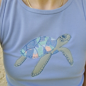 May include: A light blue ribbed sleeveless top featuring a central applique of a sea turtle. The turtle has a grey body and a shell composed of multi-colored fabric patches in shades of blue, green, and peach, outlined with dark blue stitching.